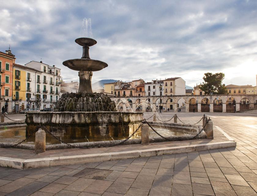 Piazza con fontana centrale, circondata da edifici storici e portici.