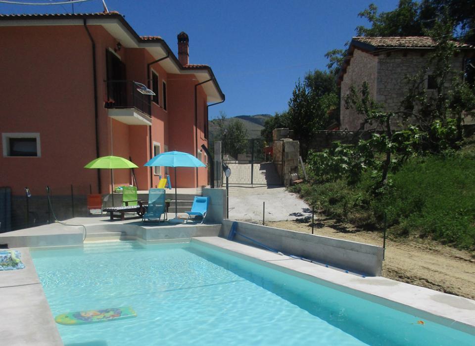 Piscina con lettini e ombrelloni accanto a una casa rosa in campagna.