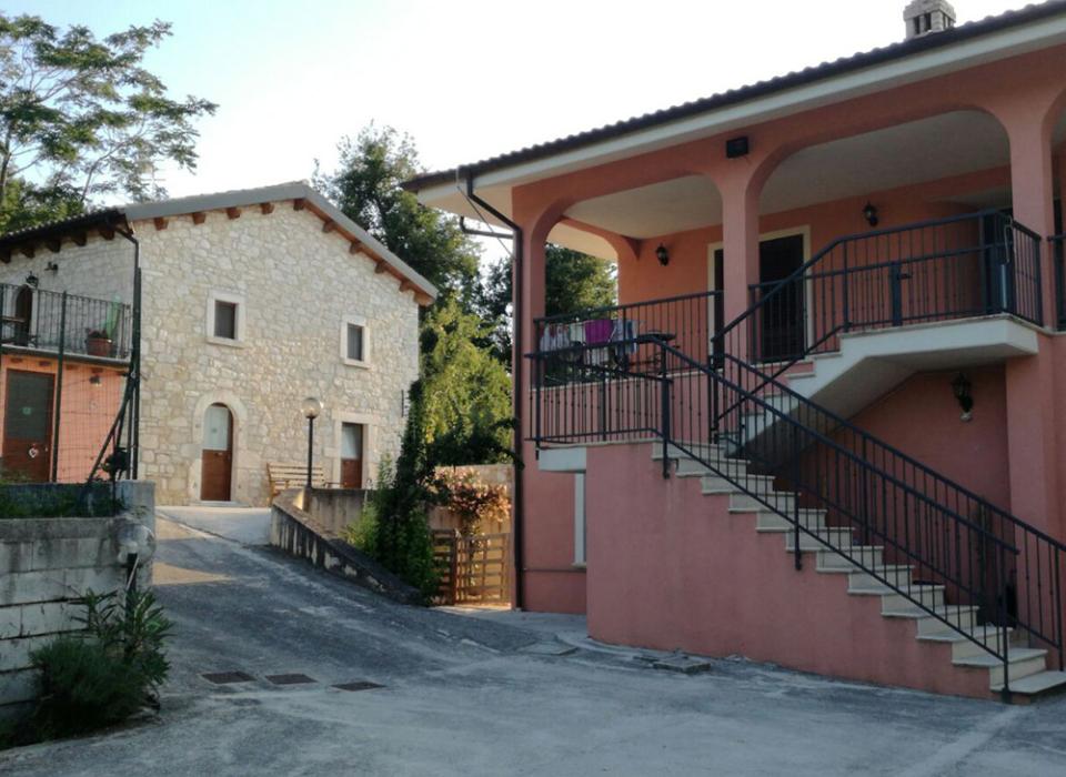 Casa in pietra e edificio rosa con scale esterne, circondati da vegetazione.